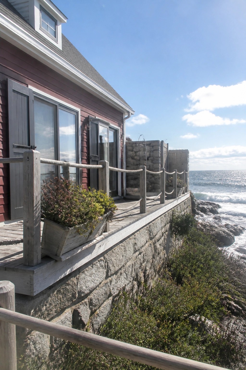 A burgundy shingled house with gray stone foundation walls, a wooden deck, and ocean views in the background.