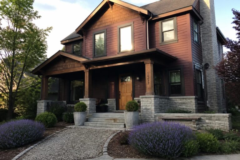 A two-story house with burgundy wood siding, gray stone foundation and porch supports, a wooden front door, and a gravel front path bordered by shrubs.