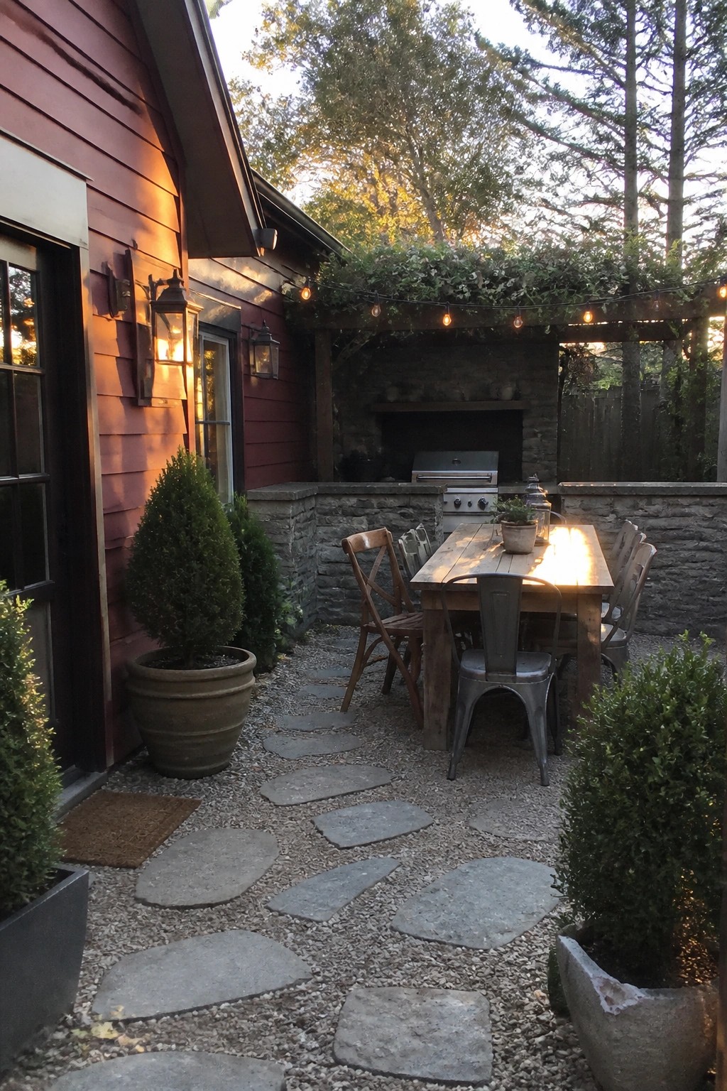 Backyard patio scene showing a wooden dining table with mixed metal and wood chairs on gravel with stone stepping pavers, beside a red house wall and a stone outdoor kitchen with built-in grill and fireplace.