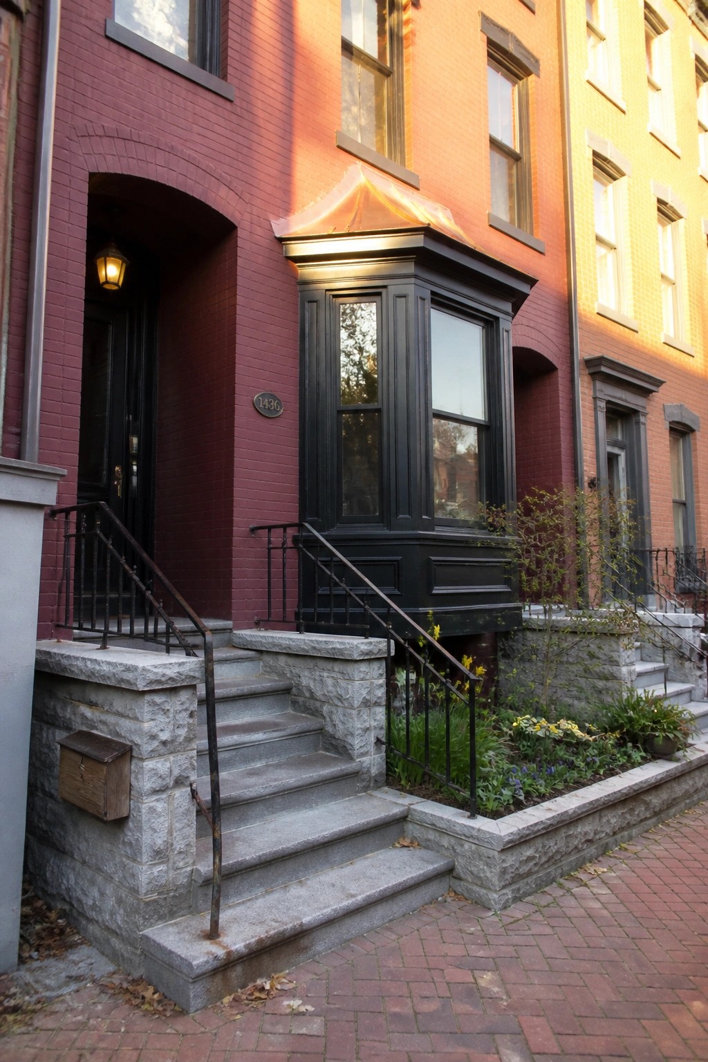 Red brick townhouse with black bay window, copper roof, stone steps, and arched doorway.