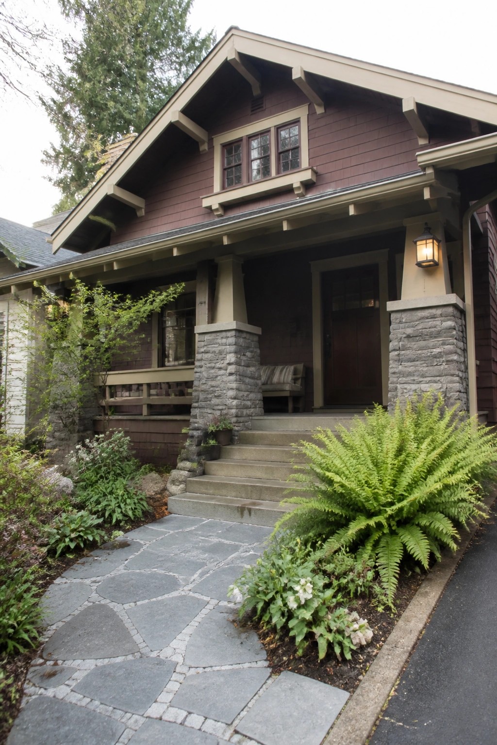 A front view of a burgundy house with gray stone pillars supporting the porch, stone steps, and a flagstone path leading to the door.