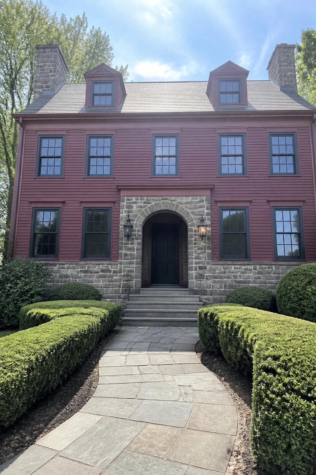 A two-story burgundy house exterior with gray stone foundation, arched stone entry around a dark door, multiple windows, and curved shrubs along a stone walkway.