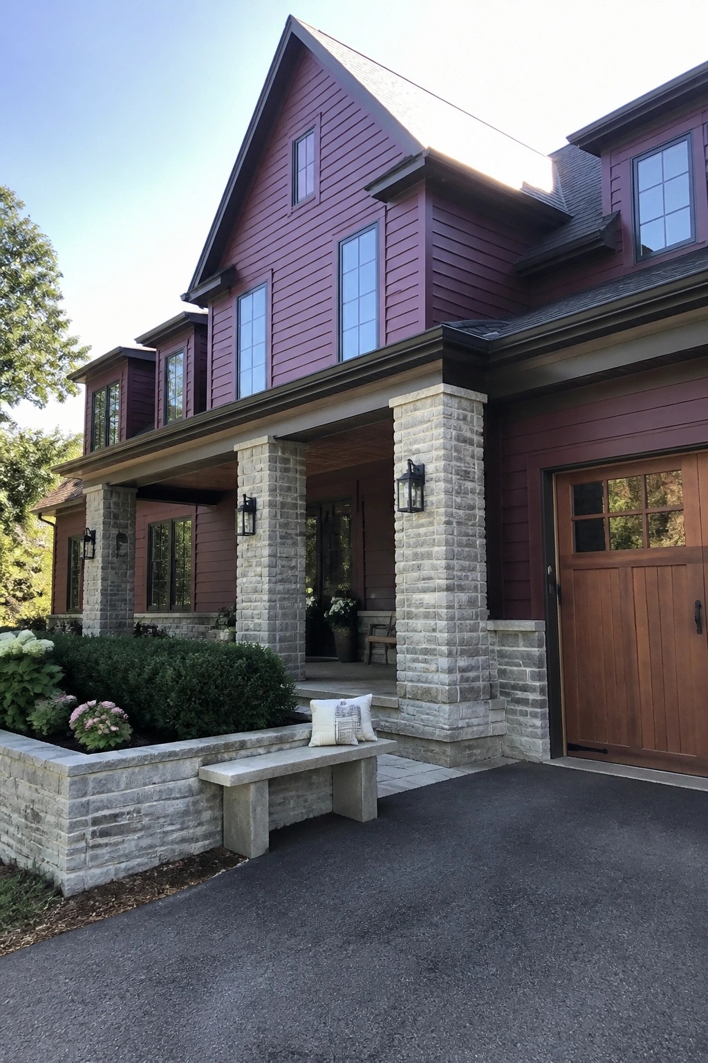 Two-story maroon house with stone porch pillars, wooden garage door, and bench with plants.
