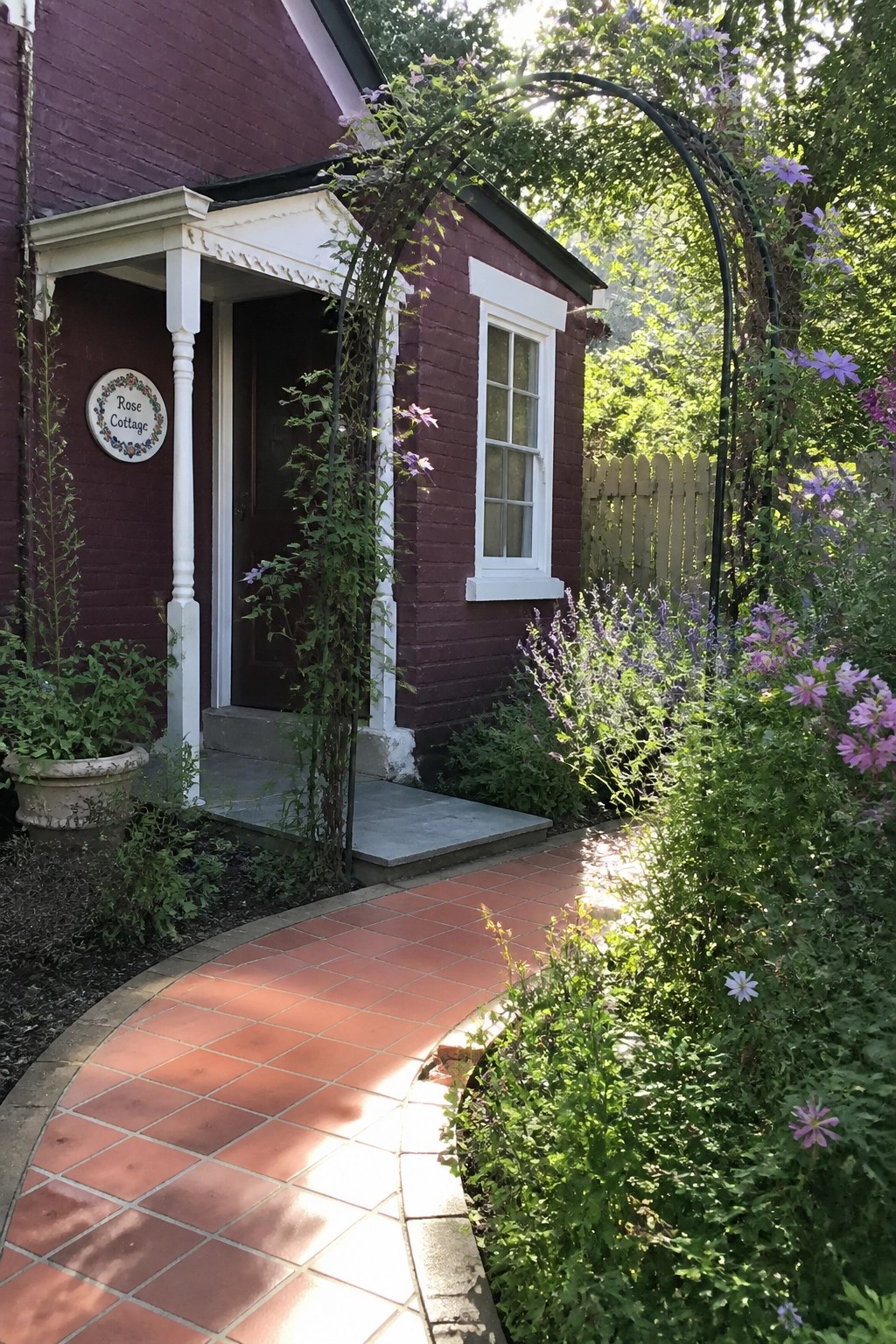 A purple brick house with white trim around the door and windows, a brick path leading to the entrance, and purple flowers growing on an arch.