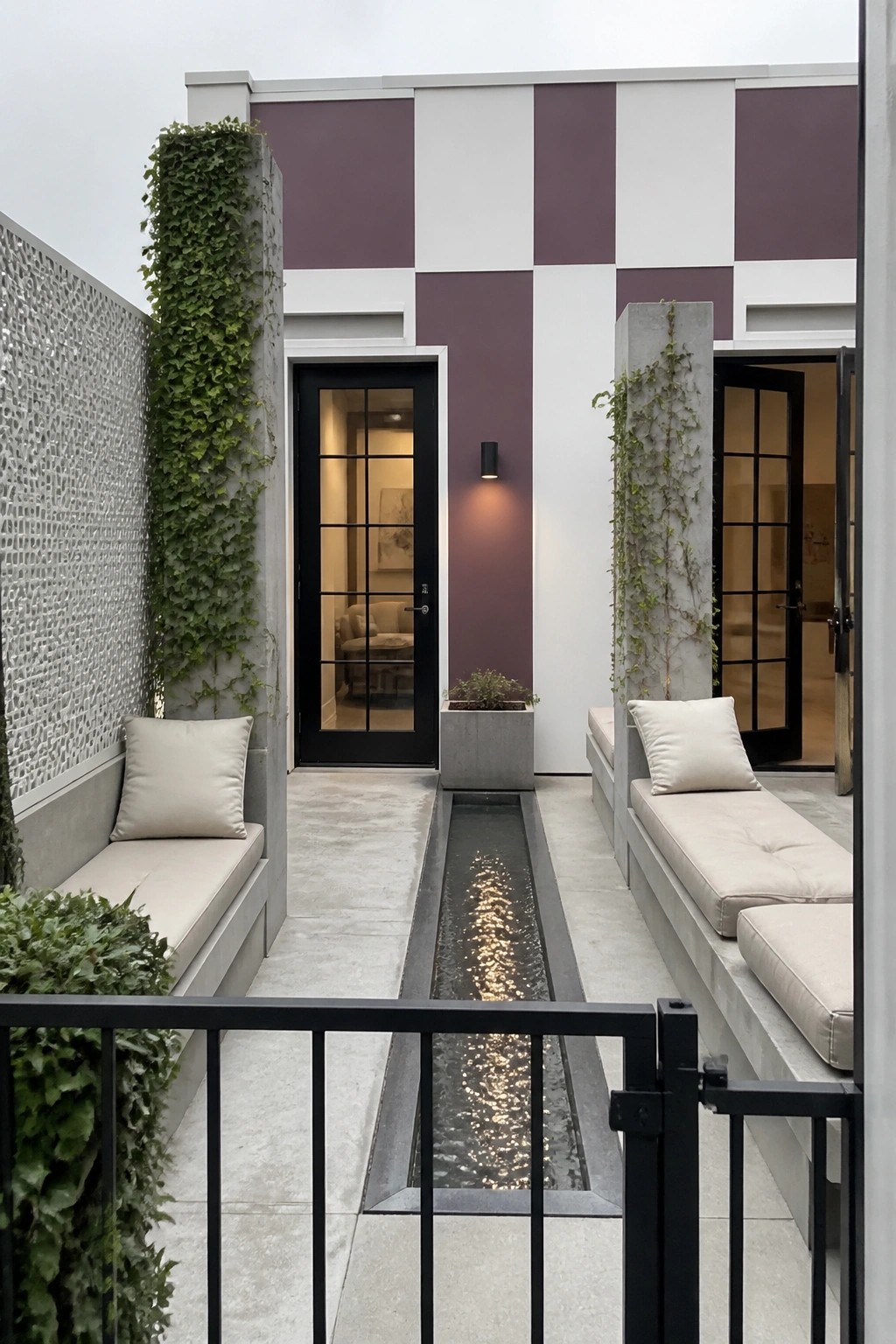 A narrow courtyard with built-in cushioned benches on both sides, a long water channel running down the center, and a rear wall painted in wide vertical purple and white stripes with black doors.
