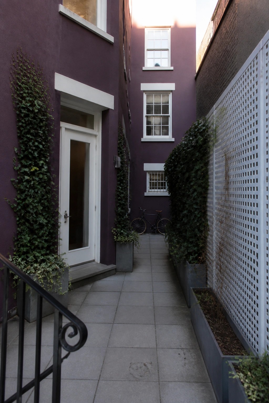 Narrow paved walkway between two buildings with deep purple walls, white door and window trim, climbing vines, and gray planters.