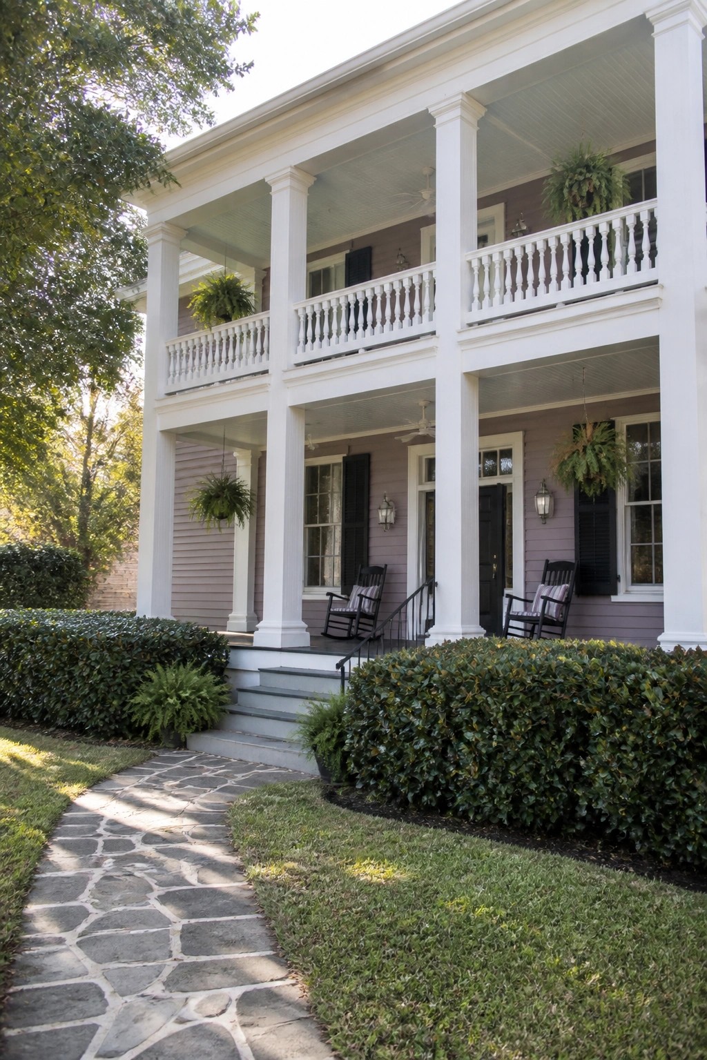 A two-story house with light purple siding, white columns, and porches, viewed from a stone pathway lined with green bushes.