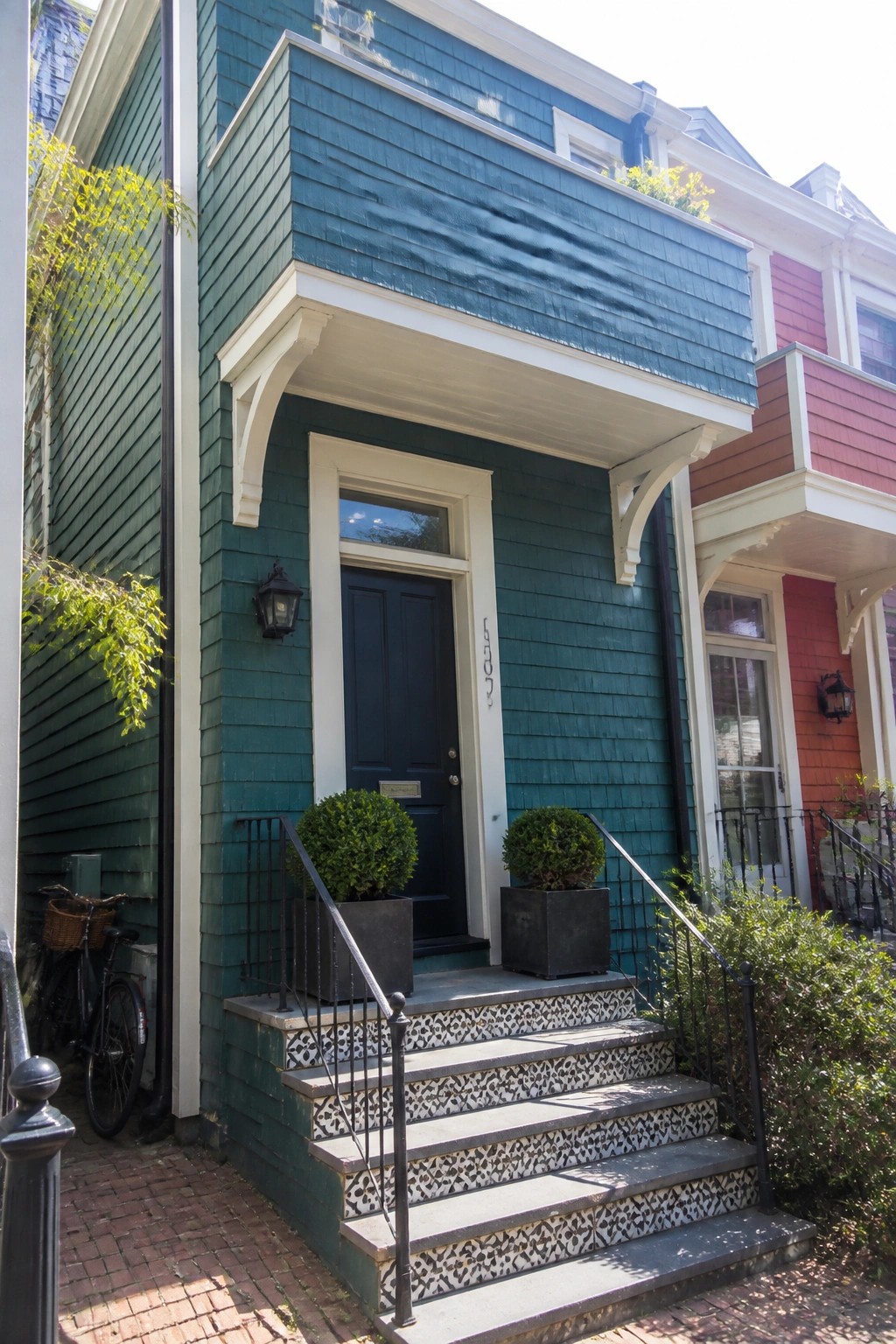 A teal cedar shake sided house with white trim around the door and windows, a dark blue front door, and tiled steps with two potted shrubs.