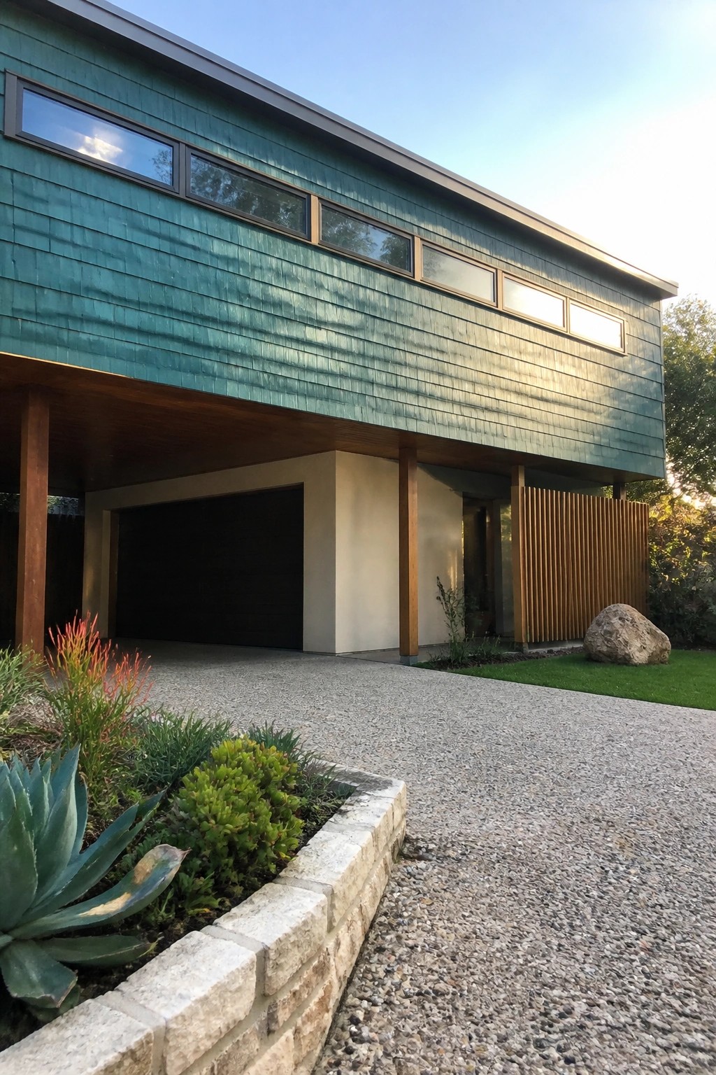 A modern two-story house with textured teal cedar shake siding on the upper level, wood support posts, and a dark garage door.