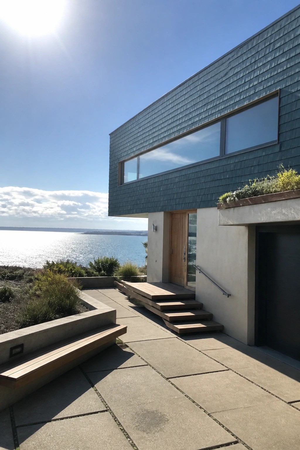 Modern house with teal cedar shake siding on the upper level, large horizontal windows, a wooden door, and concrete steps beside a body of water.