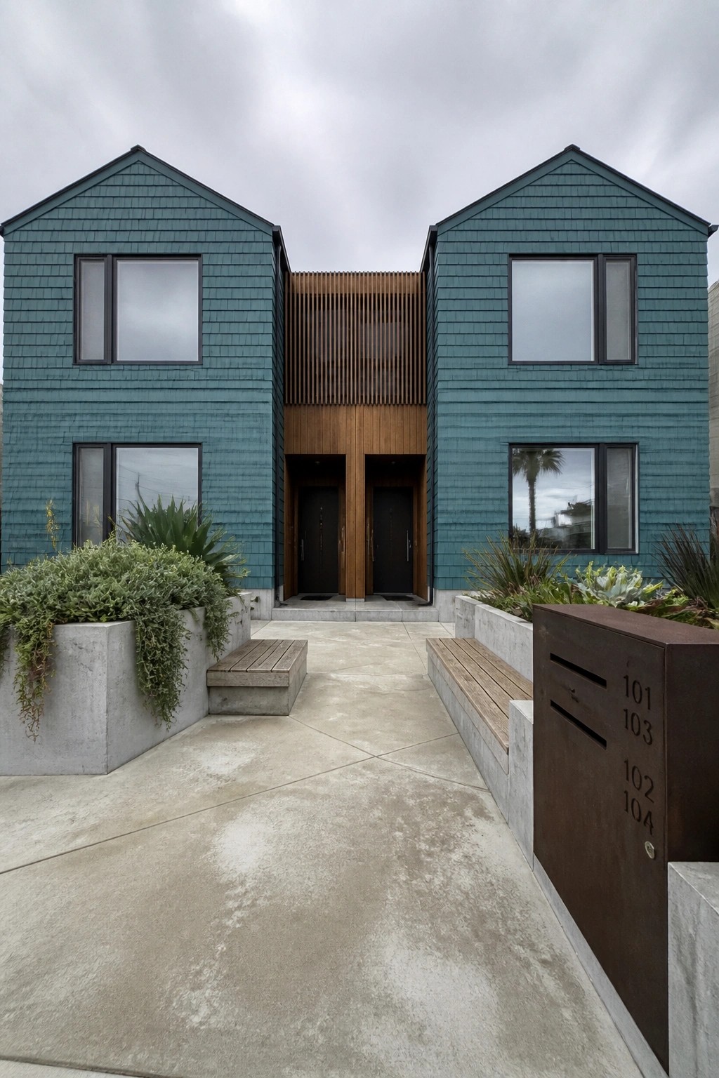 Modern teal duplex with wooden accents, concrete walkway, benches, and metal address panel.
