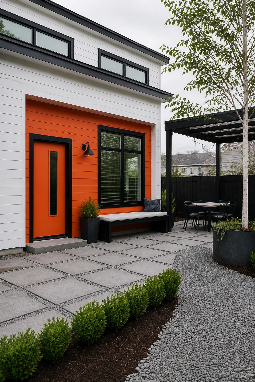 A modern house with white siding, an orange accent wall and door with black trim, a black-framed window, and a stone patio with bench, table, and gravel landscaping.