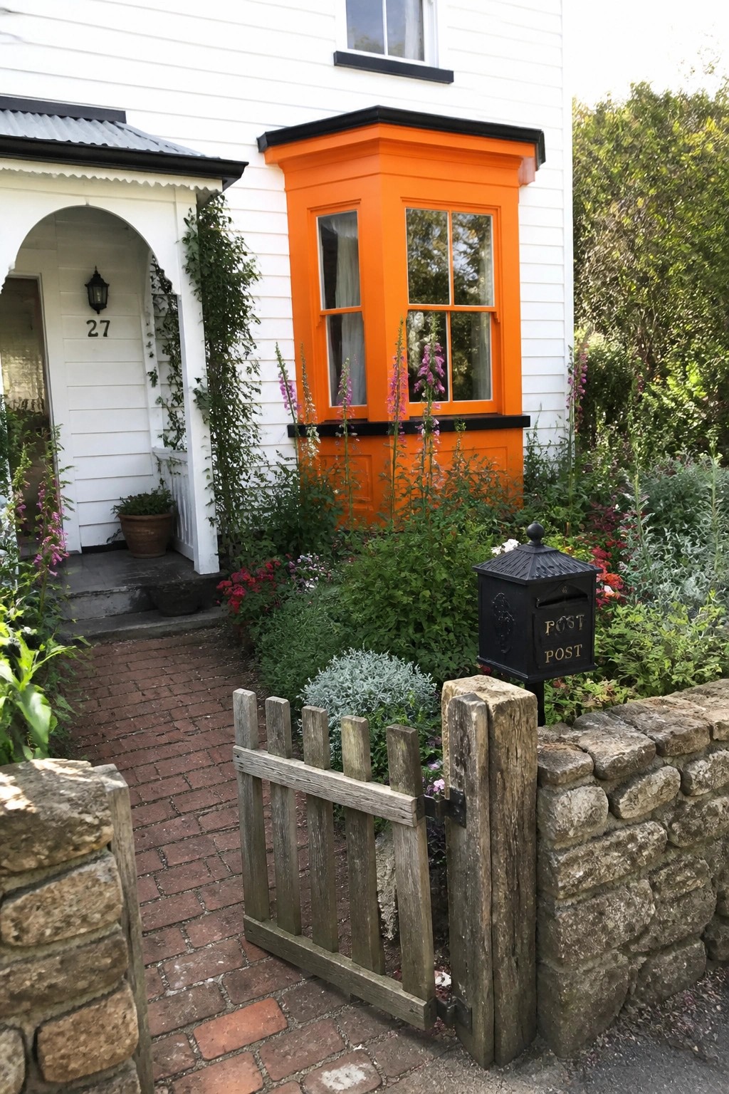 A white house with a bright orange bay window trimmed in black, next to an arched entryway, with a brick path and wooden gate in the foreground.
