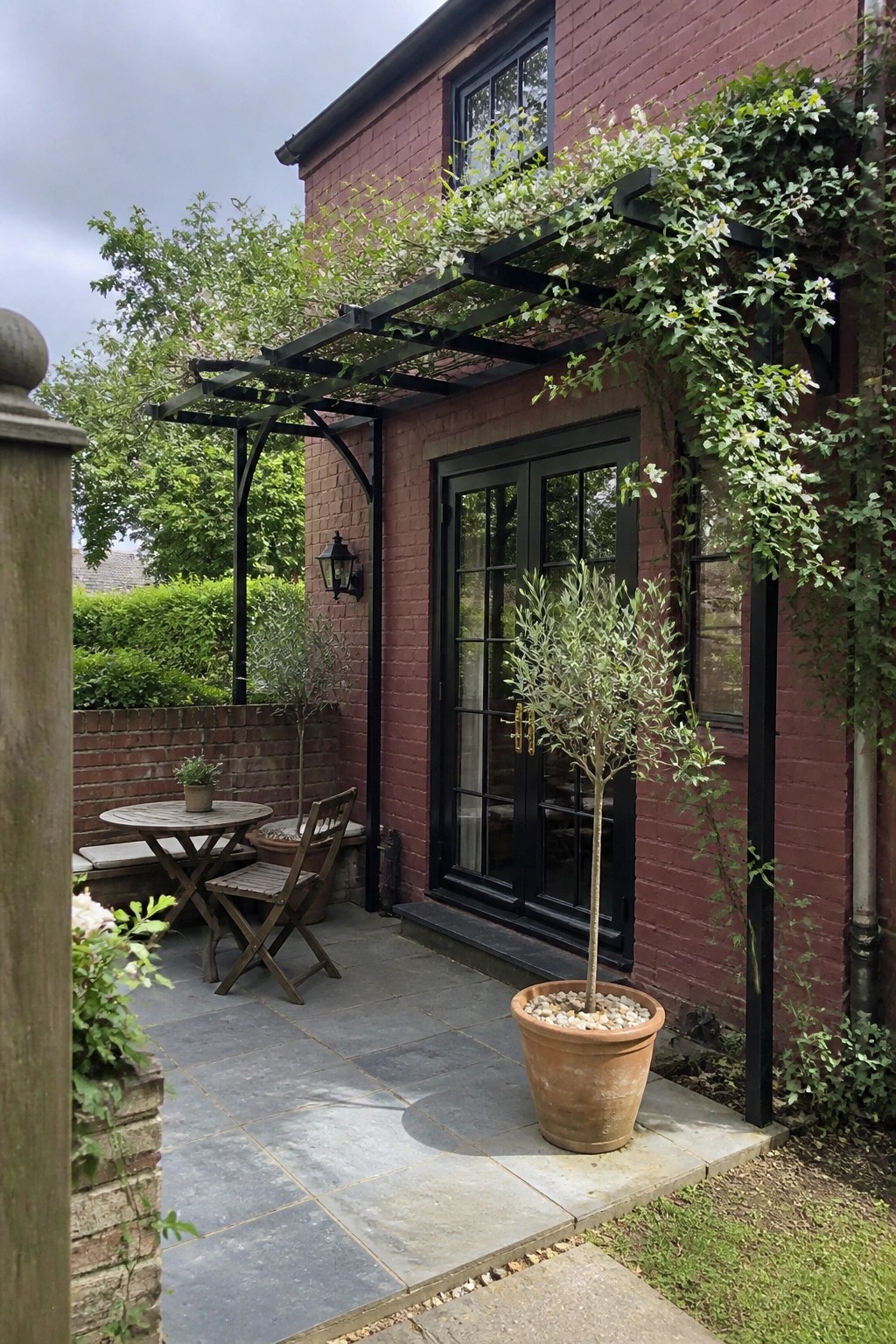 Red brick house patio with black doors, vine-covered pergola, potted olive tree, and wooden furniture.