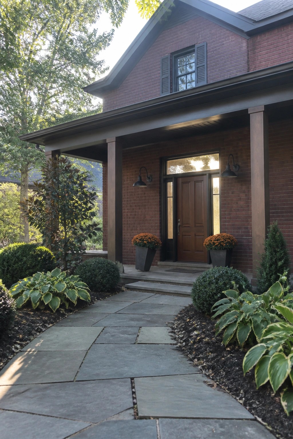 Front view of a maroon brick house with dark painted porch columns, a wooden door, black shutters, and potted plants beside stone steps.