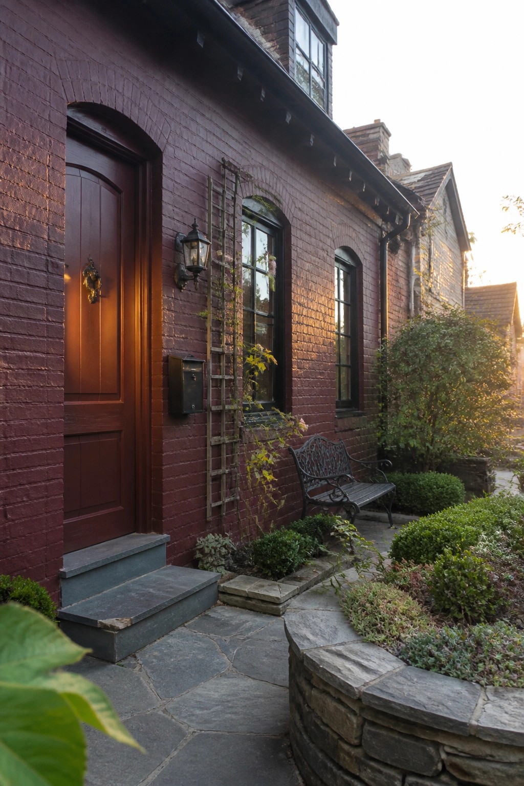 A maroon painted brick house exterior featuring an arched front door, black-framed windows, a stone path, and a small bench beside the entry.