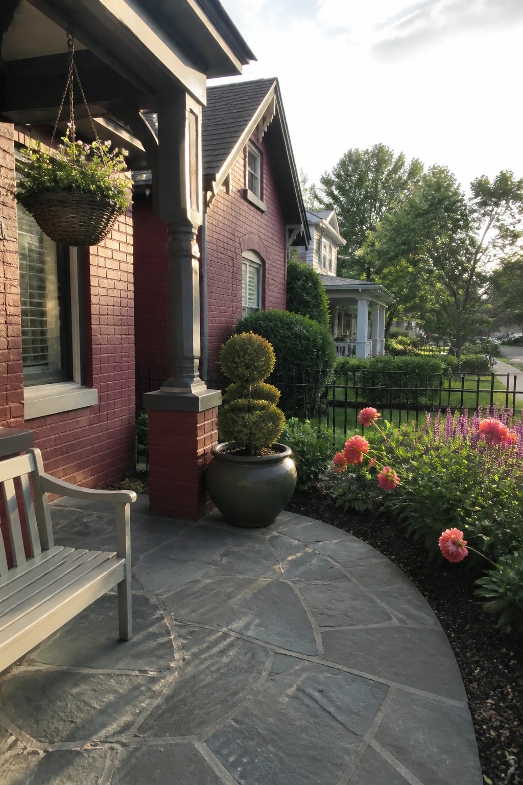 Front view of a maroon brick house with a stone porch column, gray bench, hanging basket, and curved stone patio with flowers.