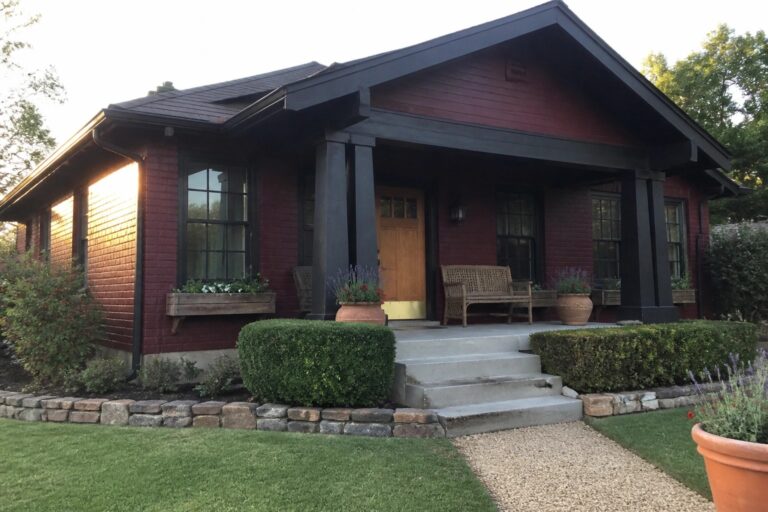 A maroon brick house exterior with dark painted trim, columns, and a gabled porch featuring a wooden front door.