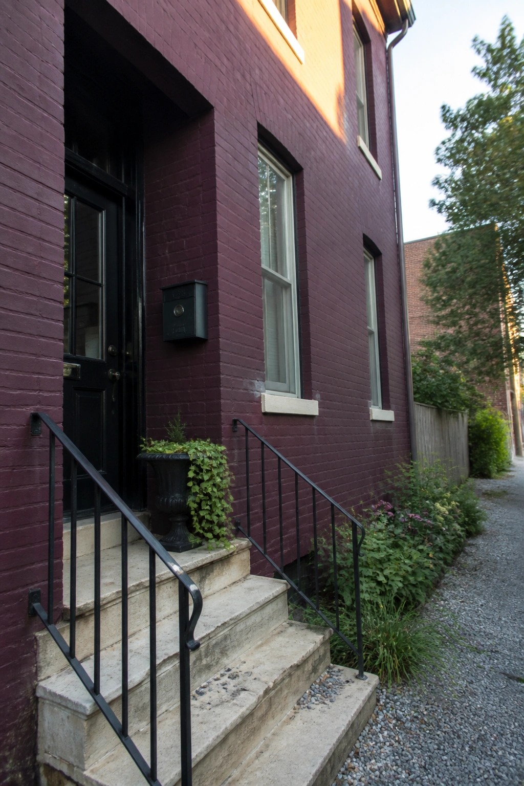 A front view of a maroon painted brick house with black door, black mailbox, black railings, and stone steps.