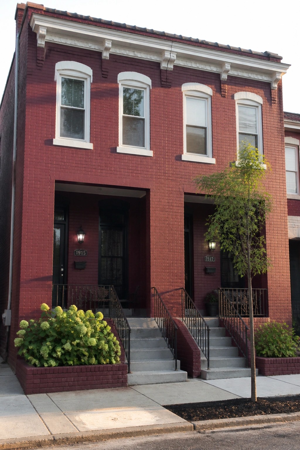 Red brick row house with dual entrances, steps, railings, and shrubs.