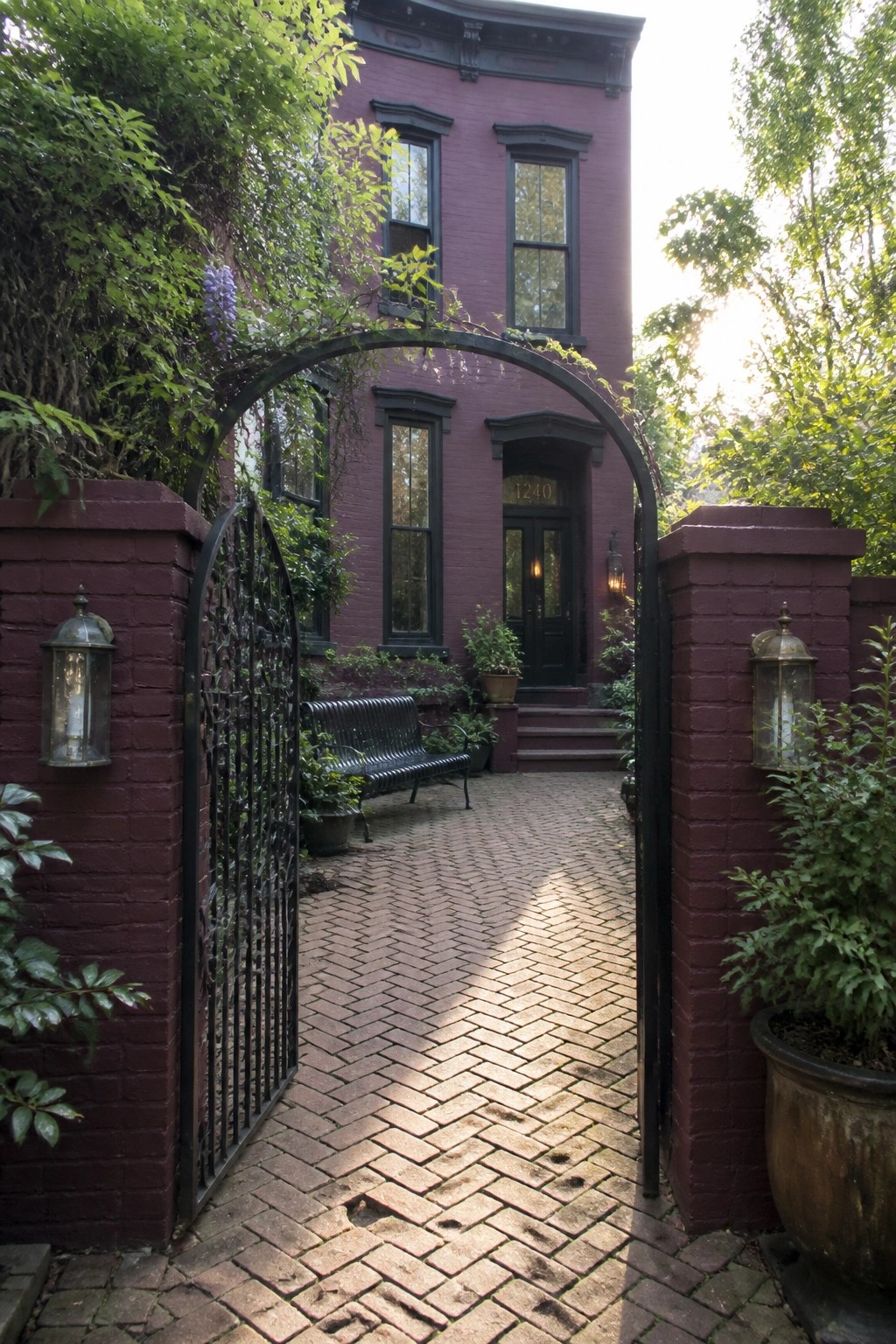 A maroon painted brick house viewed through an open black metal gate, with a herringbone brick path leading to the front door.