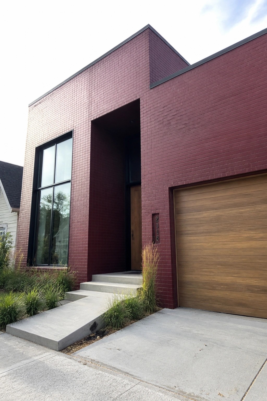 A modern two-story house with deep maroon painted brick exterior, recessed wooden front door, large black-framed window on the left, and horizontal wood garage door on the right.