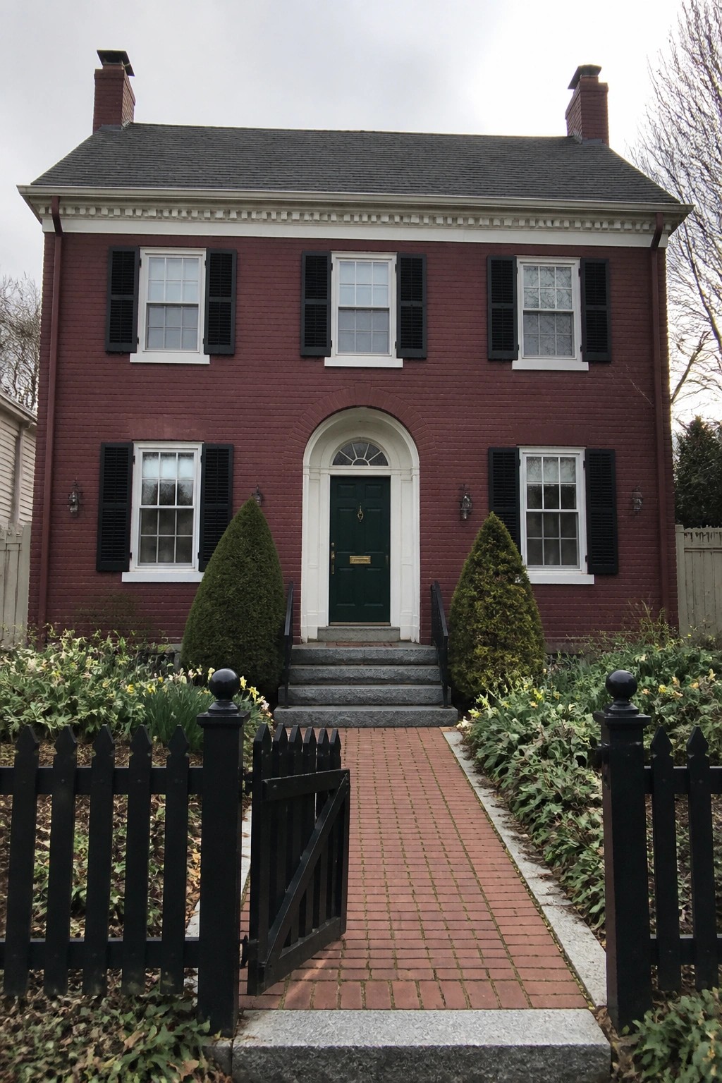 Front view of a two-story maroon brick house with white trim around the windows, an arched white entry surround, a green door, and a brick walkway leading to the steps.