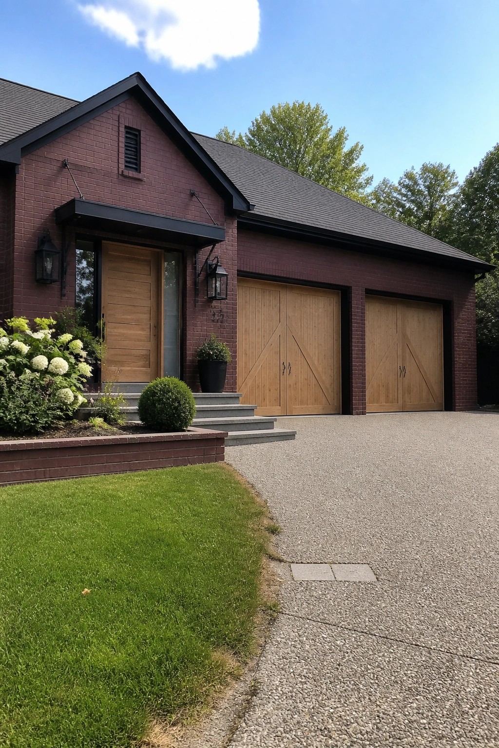 A maroon brick house exterior with black trim, a wooden front door, and two wooden garage doors facing a gravel driveway.