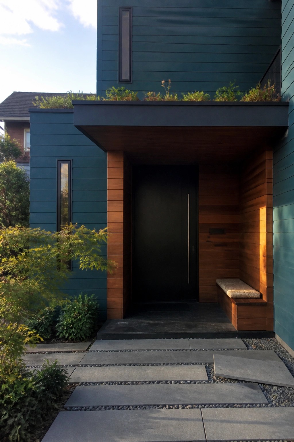 Front view of a modern house with teal siding, cedar wood panels framing a black door, and stone steps leading to the entrance.