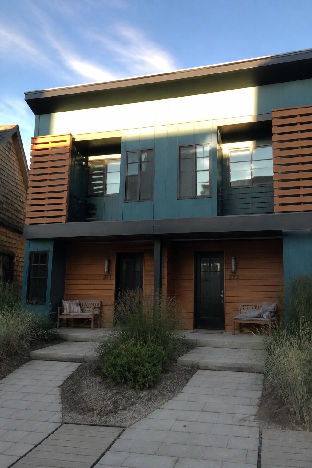 A two-story modern building with teal metal siding on the upper level and natural cedar wood on the lower level, showing two front doors and small benches.