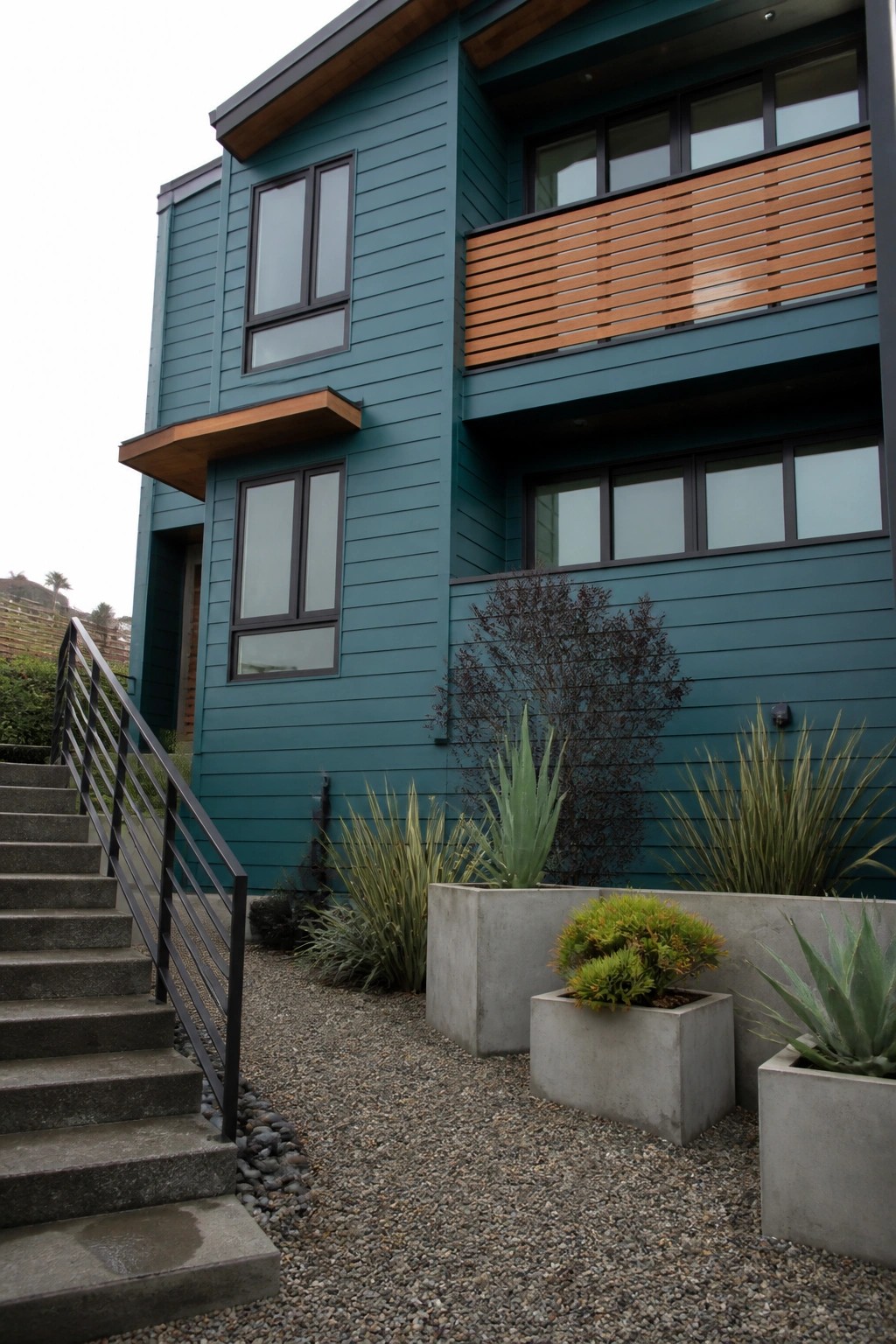 Modern teal building with wooden balcony, concrete planters, gravel yard, and stairs