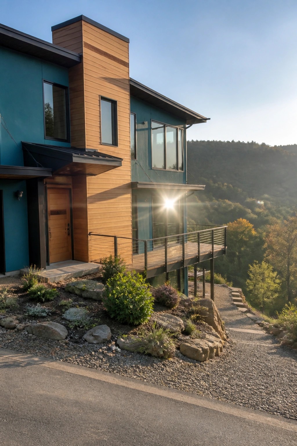 Modern teal house with wood accents, balcony, and gravel path on forested hillside