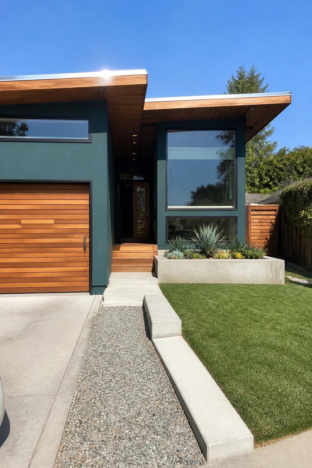A modern house with dark teal exterior walls, cedar wood accents on the roof overhang and garage door, and a concrete planter with plants beside the front walkway.