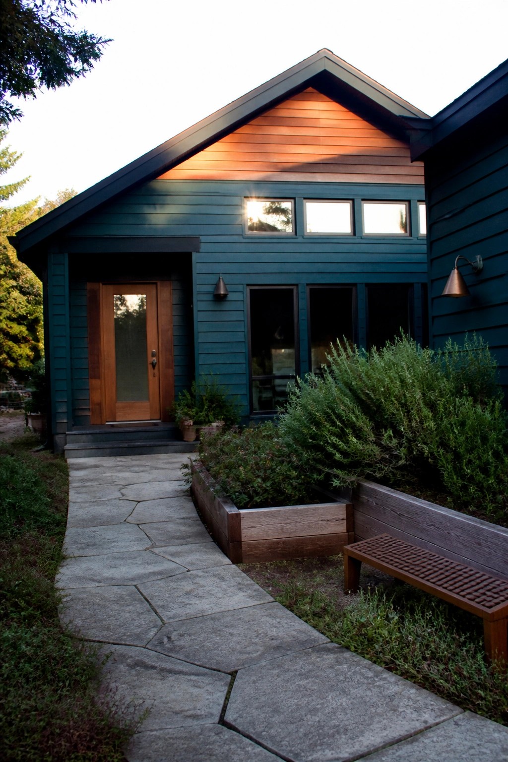 A teal house exterior with a natural wood front door, stone pathway leading to the entrance, and a wooden bench beside a raised planter.