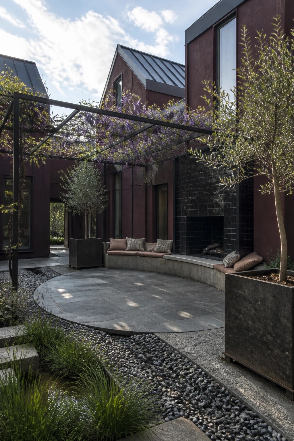 A dark burgundy house exterior with charcoal metal roof, featuring a curved concrete bench around a black tiled outdoor fireplace, a metal pergola with hanging purple flowers, and large potted trees on stone and gravel paving.