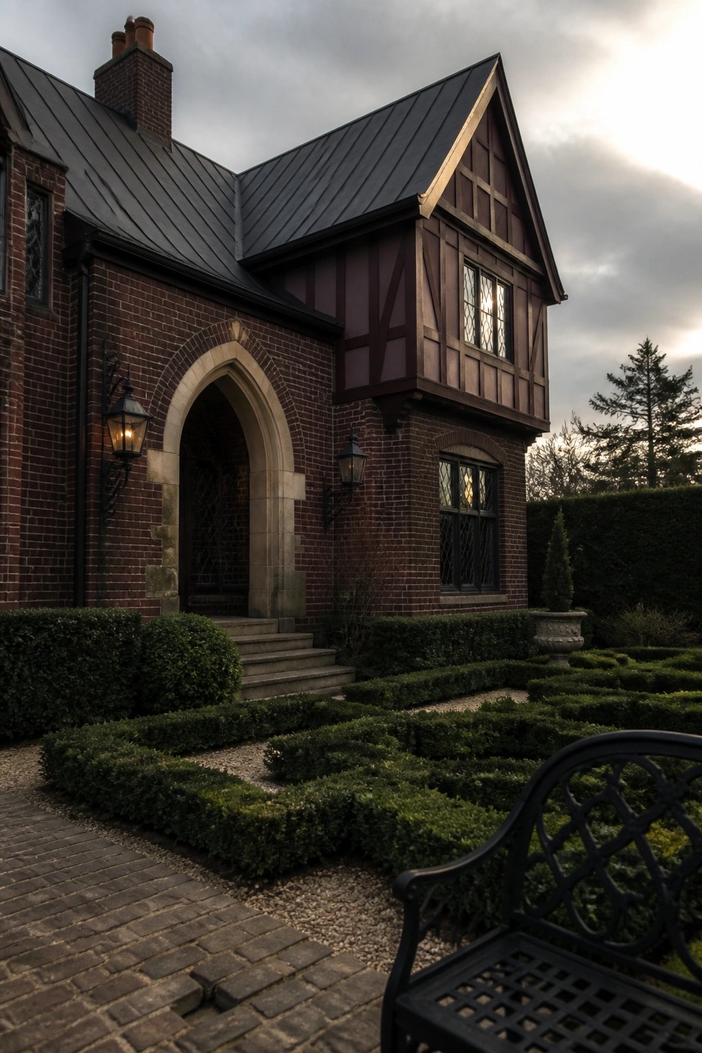 A brick house with dark burgundy half-timbering, a charcoal metal roof, and a stone arched entry next to a boxwood garden.