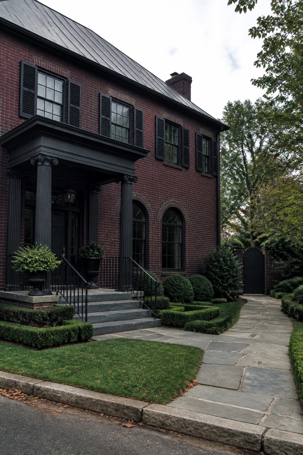 A two-story burgundy brick house with black columns, black shutters, and a charcoal metal roof, shown from the front with stone steps and a paved walkway.