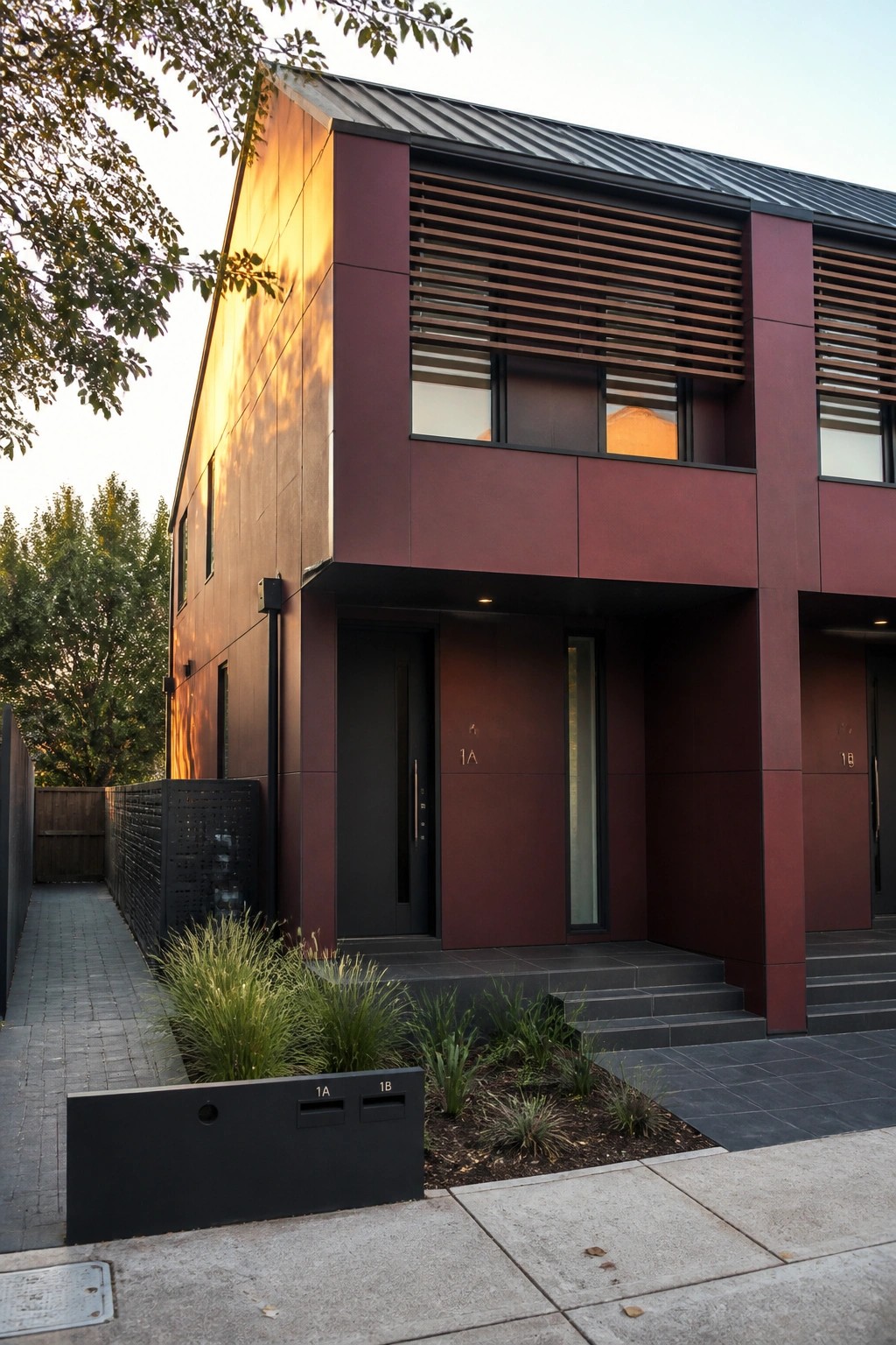 Modern maroon duplex with dark doors, wooden slats, and landscaped entrance at sunset.