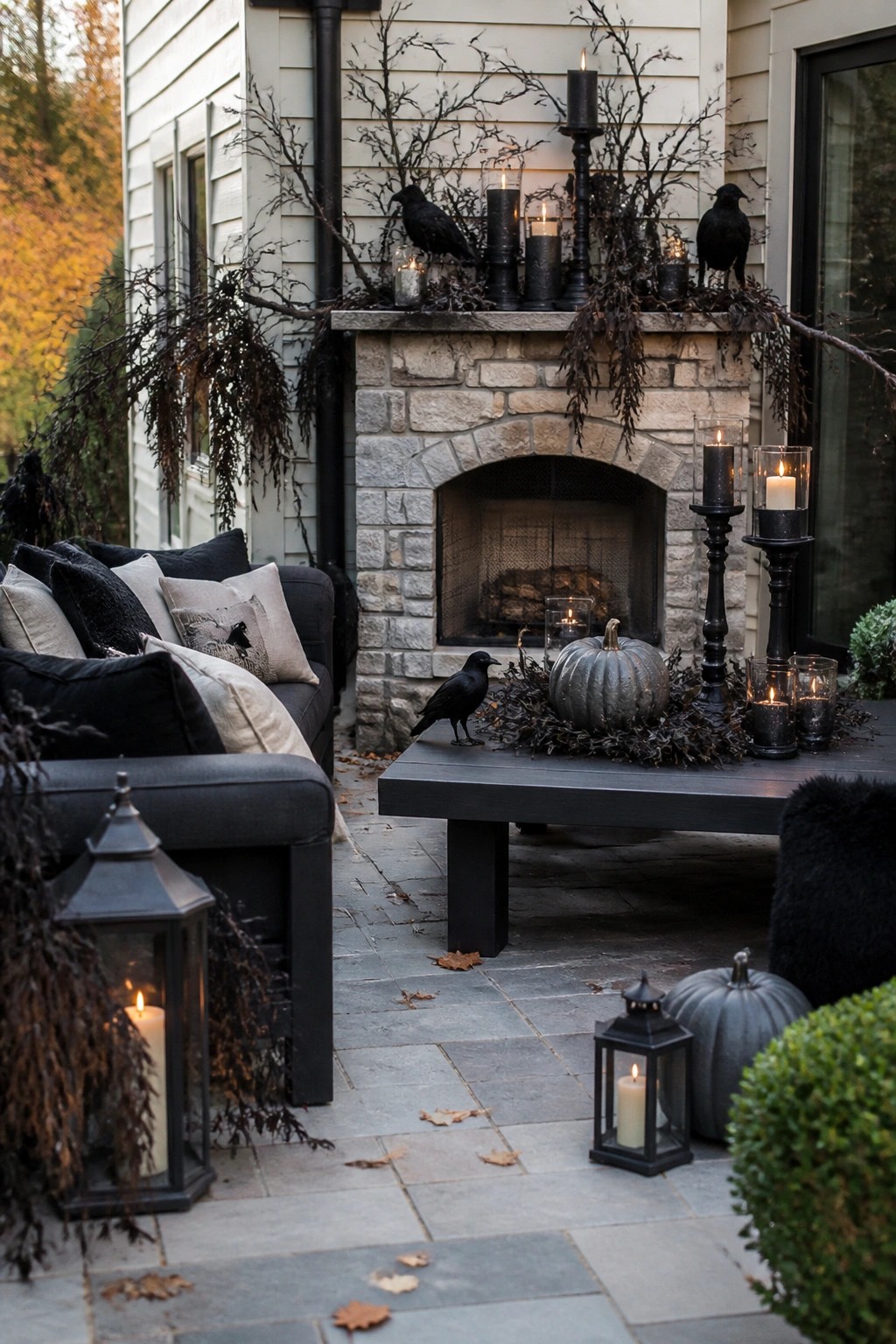 Outdoor patio with a stone fireplace decorated with black crows, black candles, and dark branches, plus a crow and gray pumpkin on a nearby table.