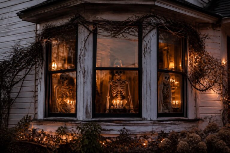 Skeleton in lace-curtained bay window with pumpkins, candles, and glowing lanterns at dusk