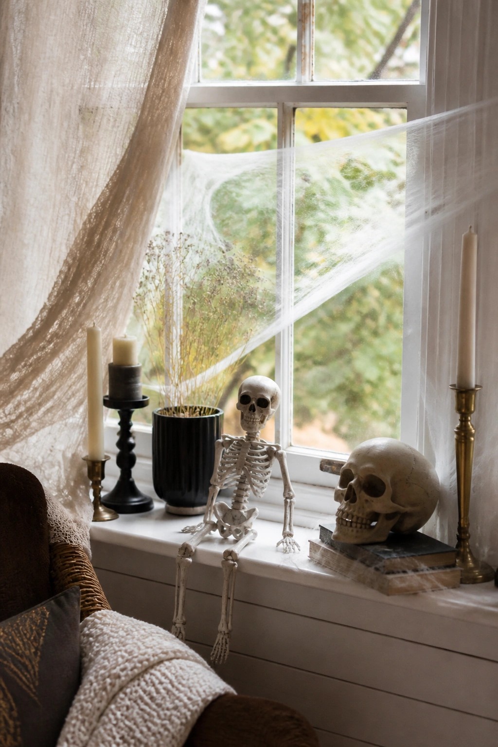 Windowsill with a small skeleton seated on the ledge, spiderwebs across the glass, a skull on books, and candles on either side.