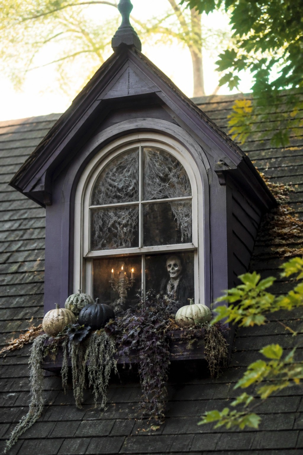 Arched purple dormer window with skeleton, candelabra, spiderwebs, pumpkins, and hanging foliage.