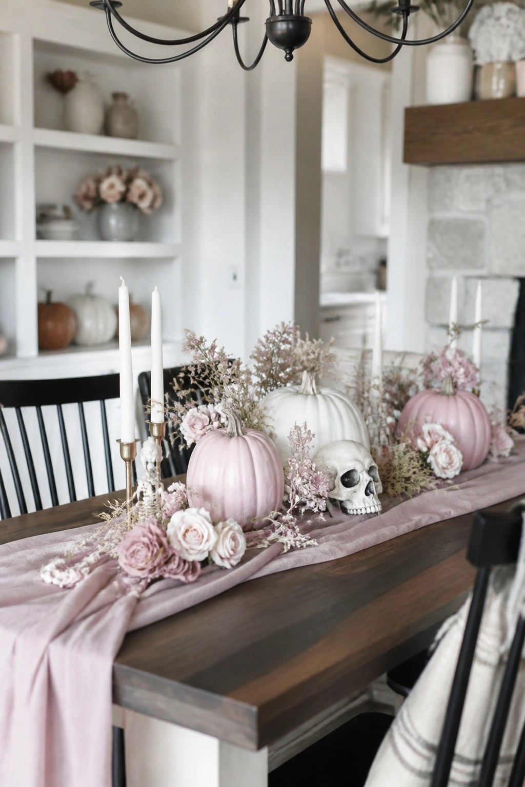 A wooden dining table decorated with a pink velvet runner, pastel pumpkins, a white skull, roses, and white taper candles.