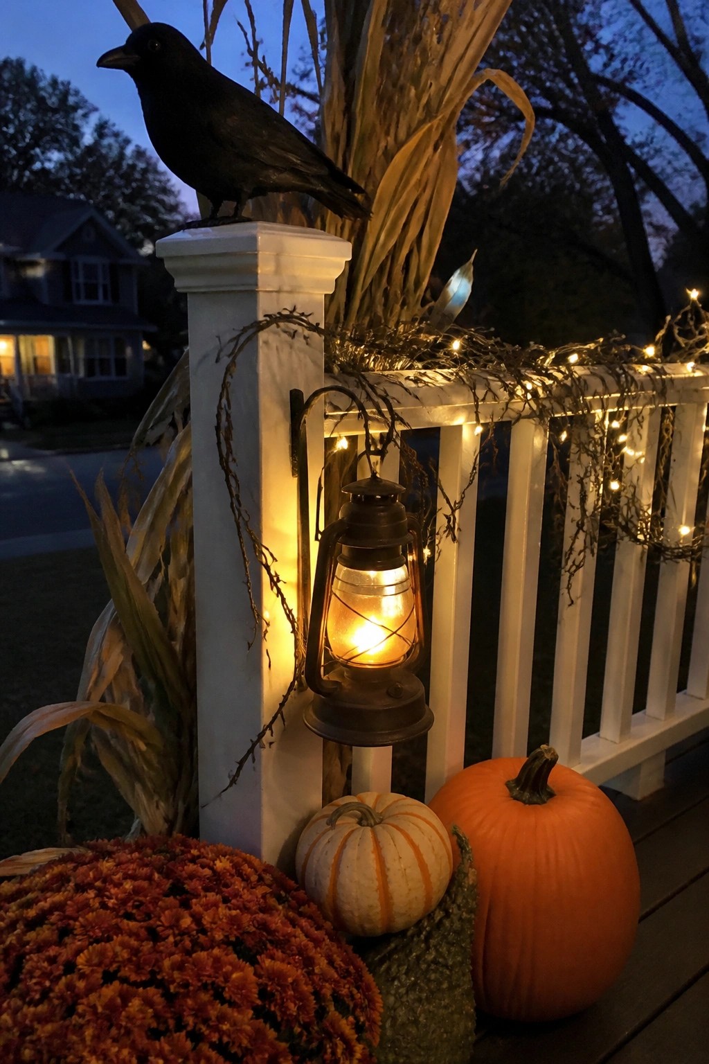 Black crow figurine perched on a white porch post beside a glowing lantern, with pumpkins and mums arranged at the base on a front porch.