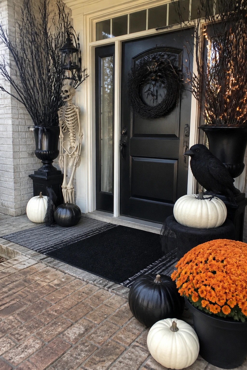 Front porch with a skeleton standing in a black urn filled with bare branches beside a black door that has a dark wreath, plus a black crow on a white pumpkin.