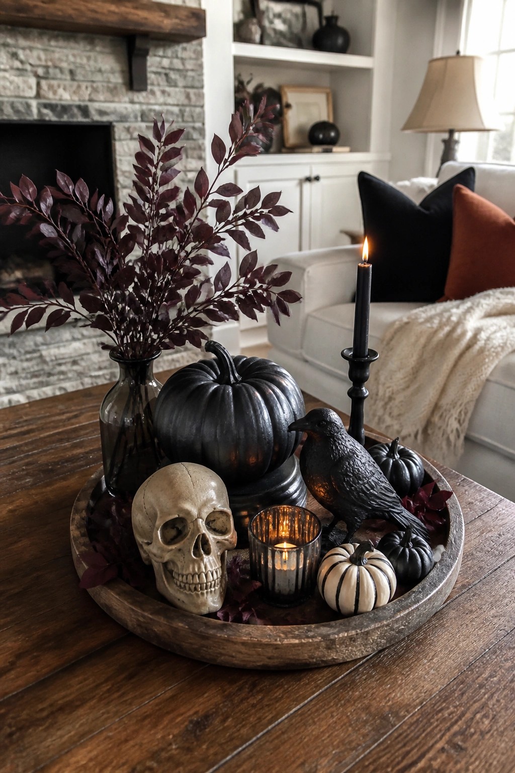 Wooden tray on a coffee table holding a skull, black crow figurine, black pumpkin, and lit candle as a Halloween display.