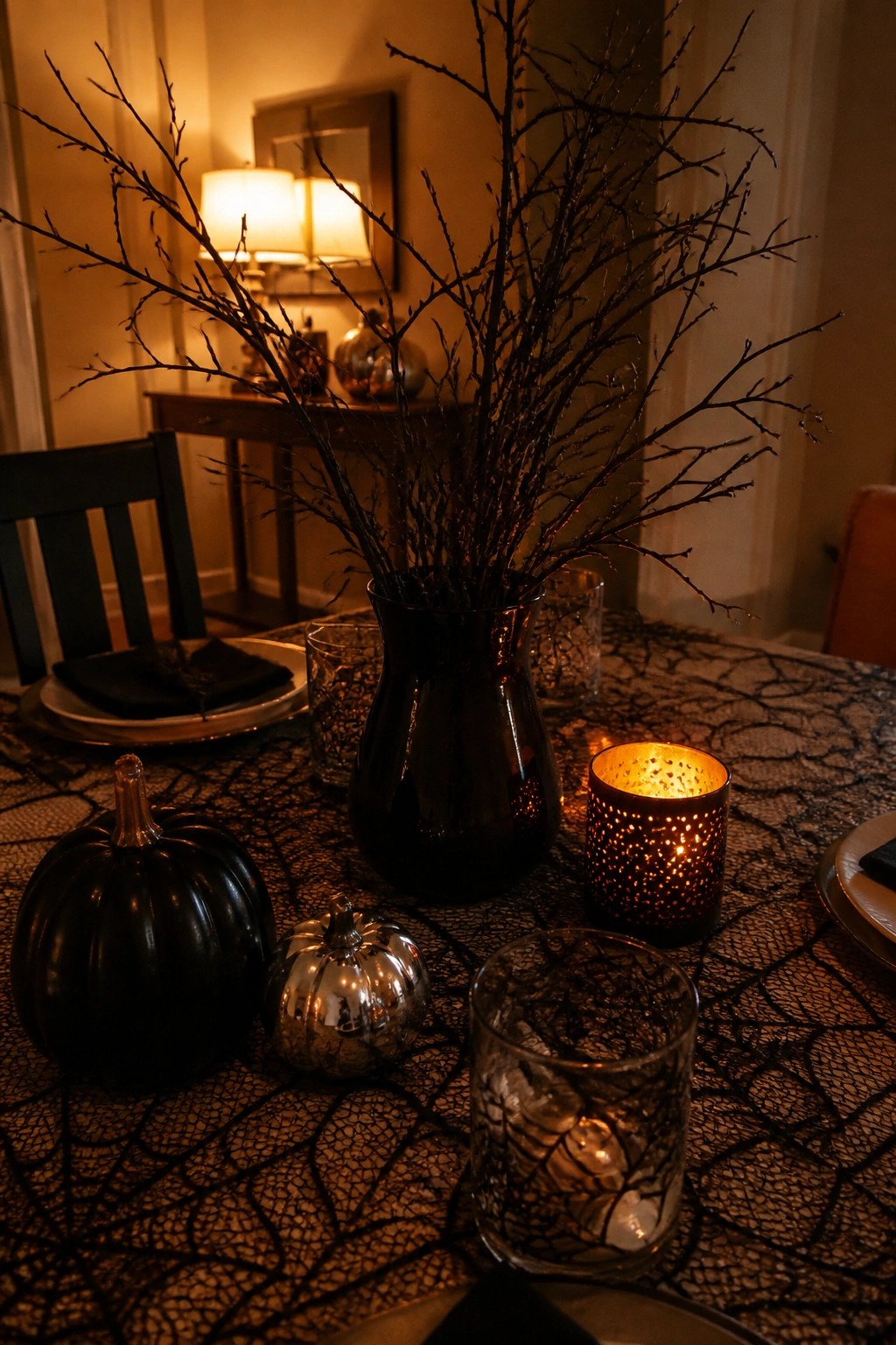 A dining table centerpiece with tall black branches in a dark vase, black and metallic pumpkins, and lit web-patterned candles on a spiderweb tablecloth.