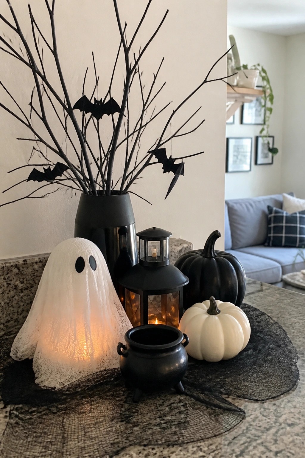 Kitchen counter vignette with a black vase holding bare branches and hanging paper bats, a glowing ghost figure, a black cauldron, lanterns, and white and black pumpkins on a lace runner.