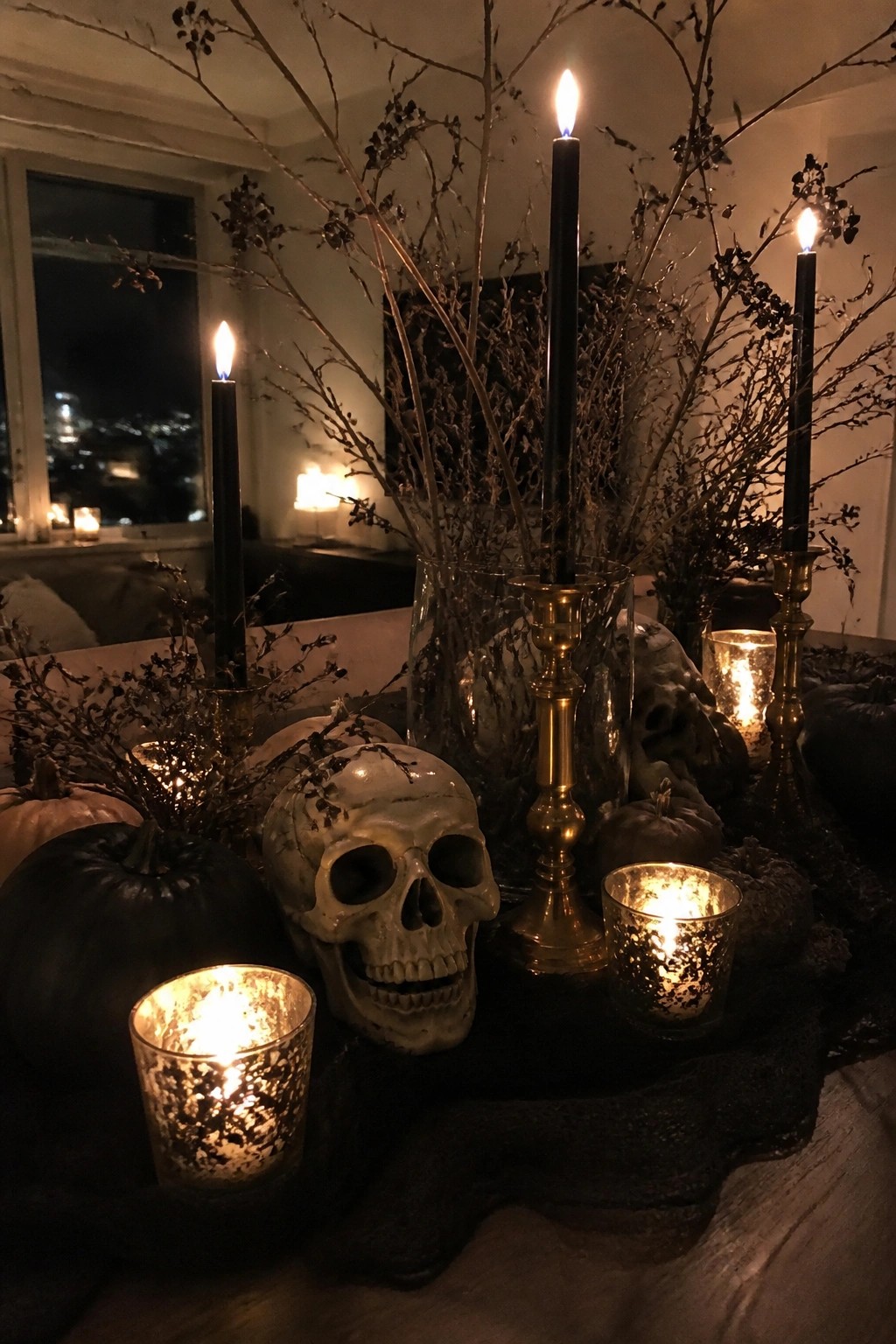 A tabletop Halloween vignette with white skulls, black taper candles in gold holders, dark branches, and black and white pumpkins arranged on a dark surface.
