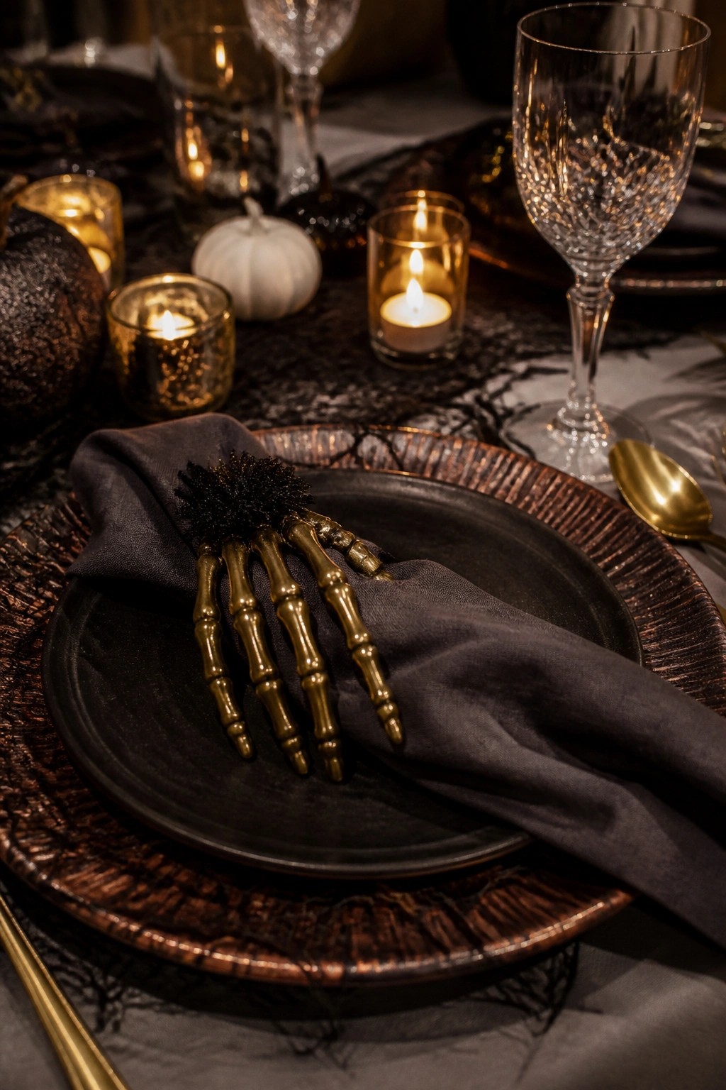 Close-up of a Halloween tablescape with a gold skeleton hand holding a black napkin on stacked dark plates surrounded by lit candles and glassware.